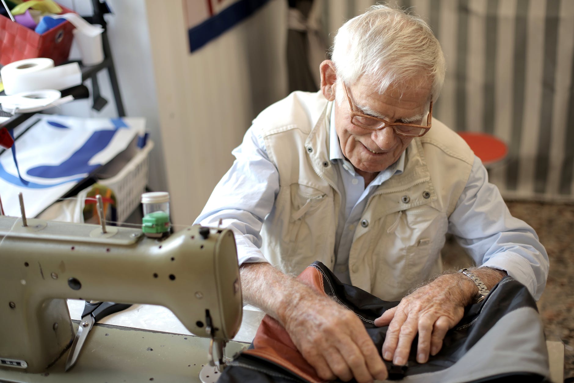 Homem idoso sorri enquanto utiliza máquina de costura que está sobre a mesa. Ele veste camisa social branca e colete creme. Ele usa um óculos de armação marrom claro. 